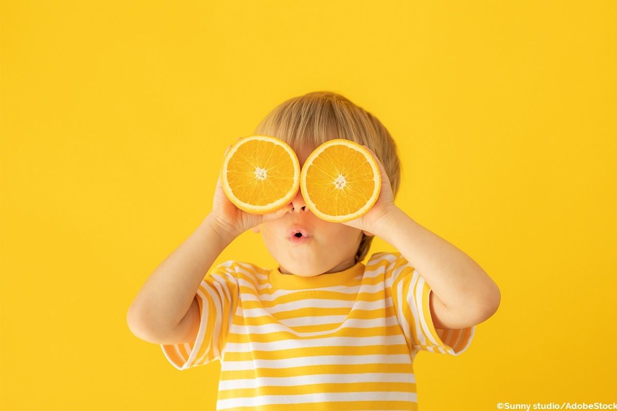 Kind hält zwei Orangenhälften vor den Augen und macht eine Grimasse.