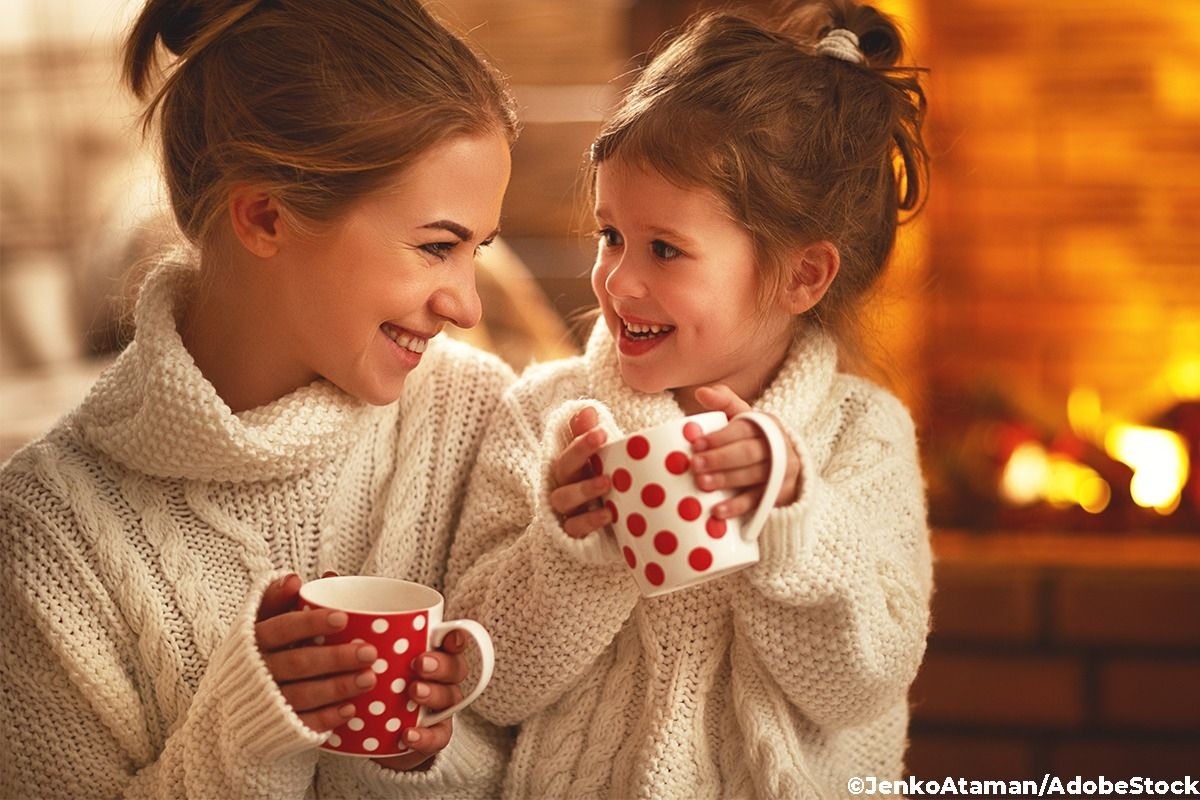 Eine Mutter sitzt mit ihrem Kind vor dem Kamin, beide halten eine Tasse hoch.