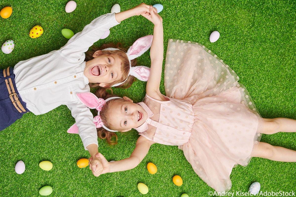 Zwei Kinder mit Hasenohrern liegen auf der Wiese, umgeben von bunten Ostereiern.