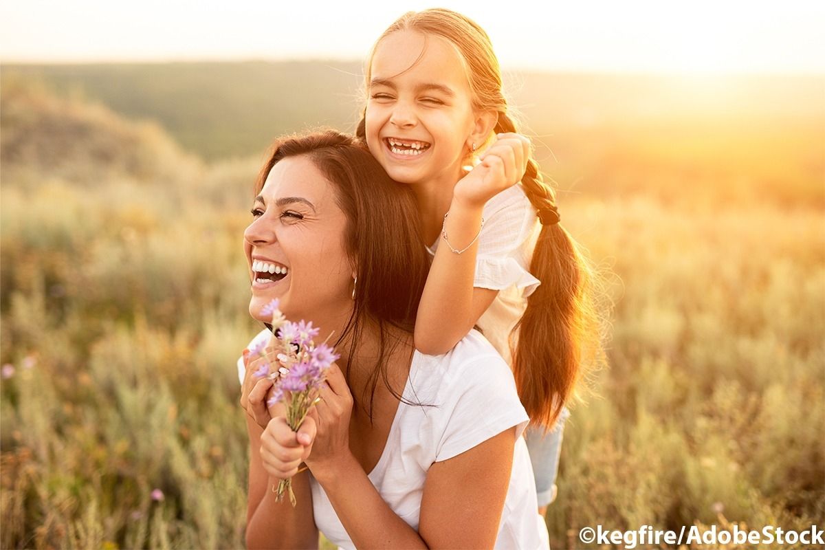 Mutter und Tochter lachen gemeinsam im Feld. Im Hintergrund ist die Abendsonne.