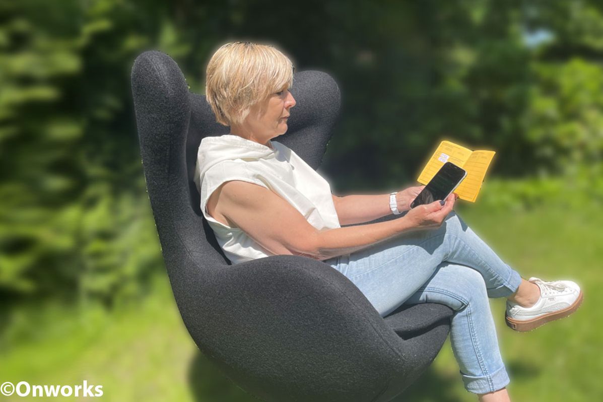 Eine Frau liest ein Buch und nutzt ein Smartphone, während sie in einem modernen Sessel sitzt