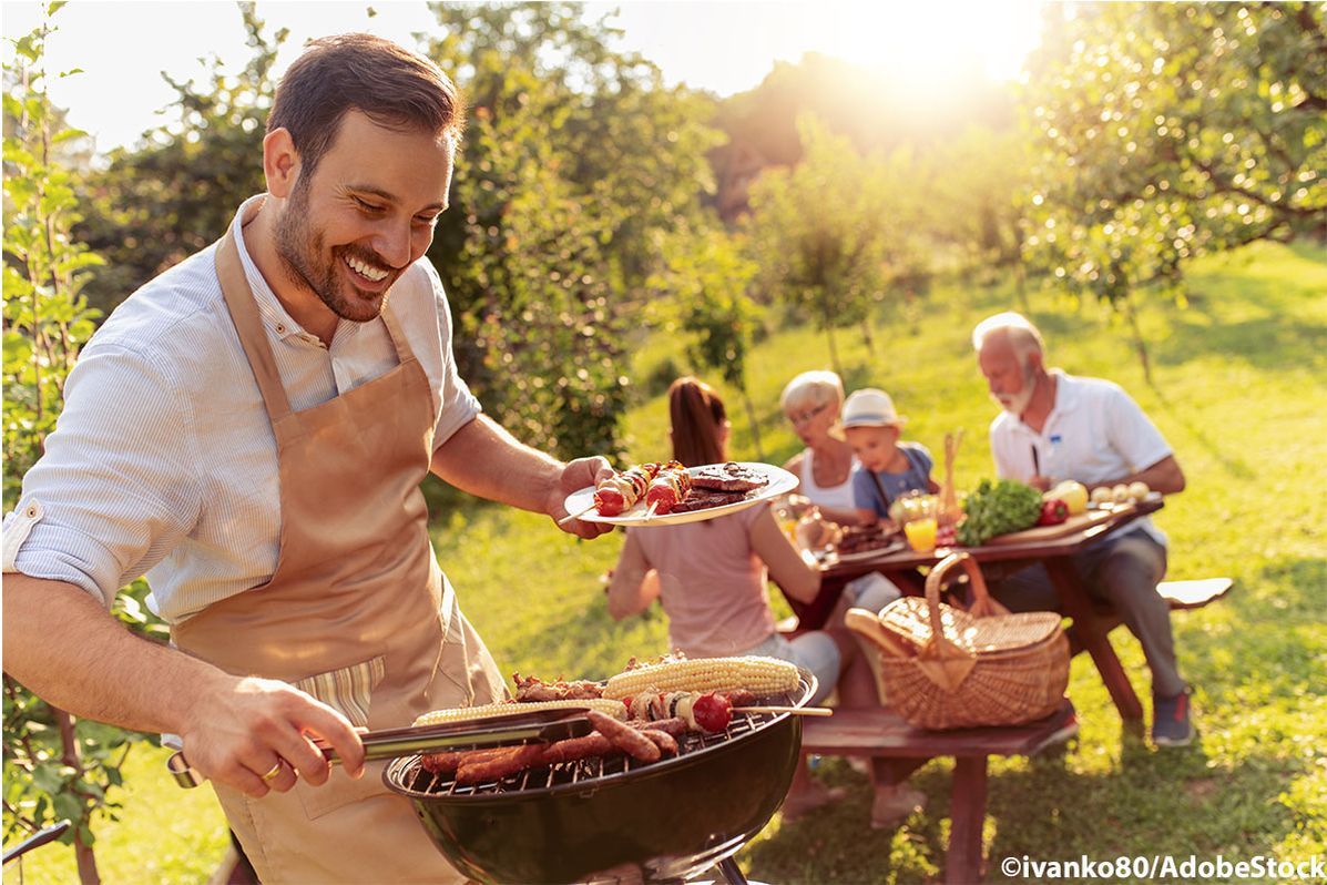 Ein lächelnder Mann grillt Würstchen und Gemüse im Freien.